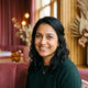 A friendly and authentic portrait of a South Asian woman smiling warmly against a soft-focus rose and amber-toned background.